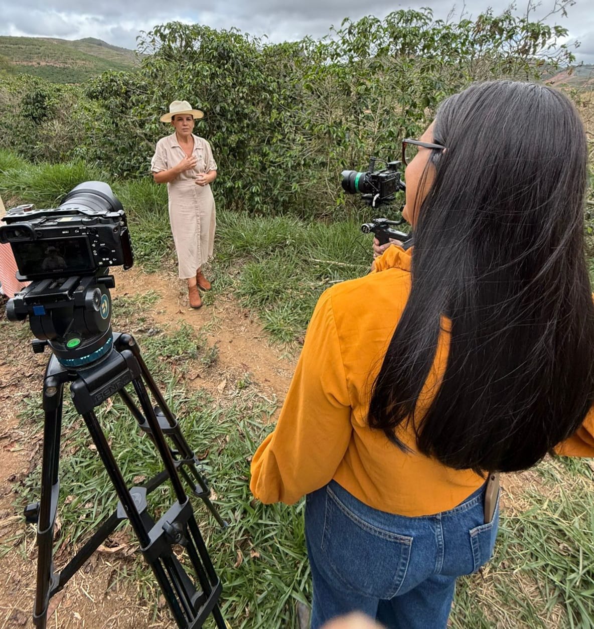 Vista dos bastidores de uma gravação de vídeo em uma área rural. Em primeiro plano, uma mulher de costas, com cabelos longos e escuros e blusa amarela, observa a cena. À esquerda, uma câmera profissional está montada em um tripé, apontada para uma mulher que está sendo entrevistada. A entrevistada veste um vestido bege, chapéu de palha e botas, posicionada em um caminho de terra entre pés de café verdes. O céu está nublado e o terreno é inclinado.