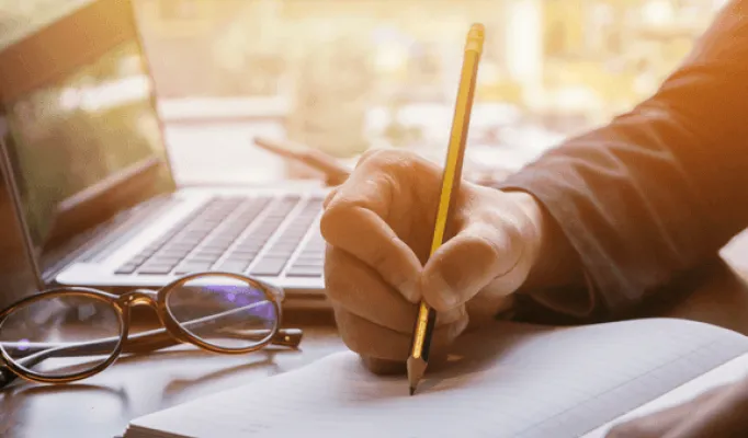 Imagem mostra uma mão com um lápis escrevendo em um caderno