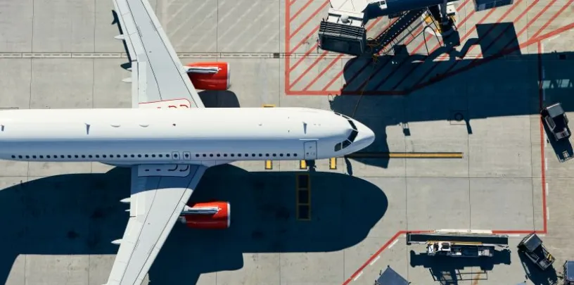 Vista aérea de avião estacionado no aeroporto, conectado à ponte de embarque e cercado por equipamentos de solo.