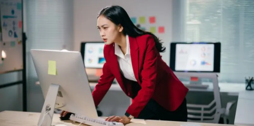 Pessoa em ambiente de escritório utilizando um computador desktop, inclinada sobre a mesa com as mãos apoiadas no teclado e no tampo. Ao fundo há outros monitores com gráficos e post-its coloridos na parede, além de documentos e materiais espalhados, sugerindo análise de dados e trabalho intensivo.