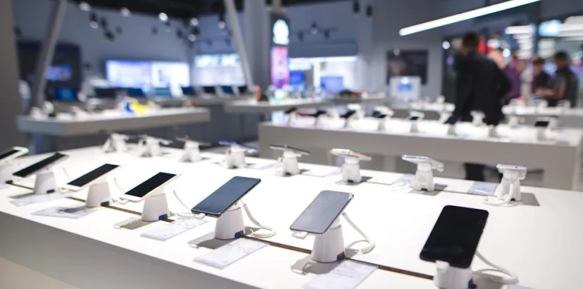 Exposição de smartphones alinhados sobre balcões em loja de eletrônicos, com dispositivos conectados a suportes de segurança e etiquetas de preço