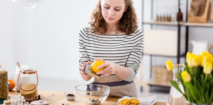 Pessoa em uma cozinha clara utilizando um ralador para raspar casca de laranja sobre uma tigela de vidro em cima da bancada. Ao redor há ingredientes e utensílios culinários, como potes de temperos, colheres, fatias de laranja e um vaso com flores amarelas, sugerindo preparo de receita caseira.