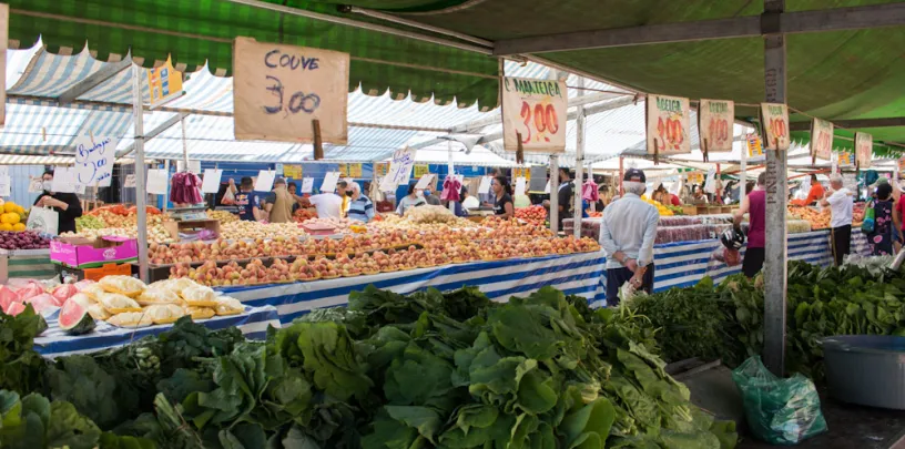 Bancas de frutas e verduras em feira ao ar livre, com produtos organizados sobre mesas e placas de preços visíveis, como ‘couve 3,00