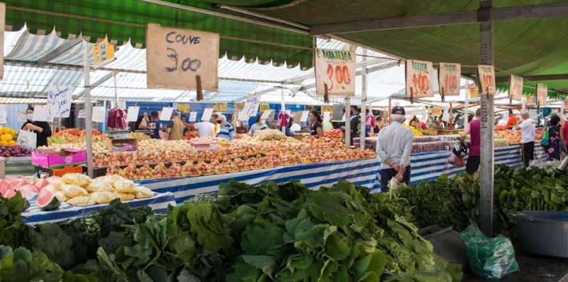 Bancas de frutas e verduras em feira ao ar livre, com produtos organizados sobre mesas e placas de preços visíveis, como ‘couve 3,00