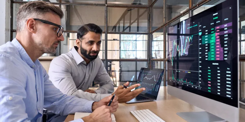 Duas pessoas sentadas em uma mesa de escritório analisam gráficos financeiros exibidos em um monitor grande, enquanto utilizam um laptop e fazem anotações. A tela mostra dados de mercado com linhas e indicadores, sugerindo análise de investimentos e tomada de decisão em ambiente corporativo.
