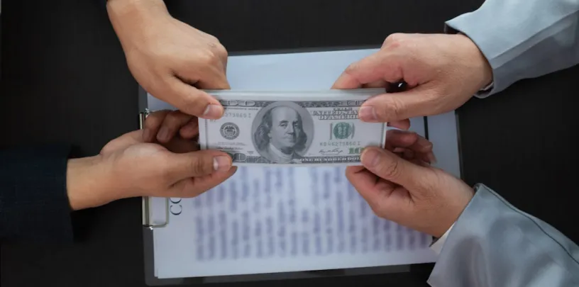 Quatro mãos de pessoas vestindo roupas formais seguram simultaneamente uma nota de 100 dólares sobre uma prancheta com documentos desfocados ao fundo. A cena sugere negociação financeira, disputa ou possível situação de suborno em ambiente corporativo.