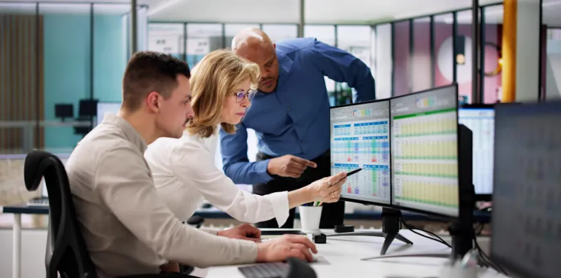 Três profissionais analisam dados em monitores de computador, apontando para planilhas e gráficos em ambiente de escritório colaborativo
