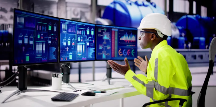 Pessoa usando capacete de segurança e roupa de alta visibilidade, sentada diante de vários monitores com gráficos e diagramas industriais. As telas exibem dados de processos, enquanto a pessoa interage gesticulando, em um ambiente que parece uma planta industrial com equipamentos ao fundo.