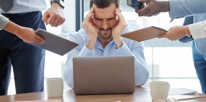 Pessoa sentada à mesa de escritório em frente a um laptop, com as mãos nas laterais da cabeça, enquanto outras pessoas ao redor estendem dispositivos como celular, tablet e bloco de anotações em sua direção. A mesa possui uma xícara e objetos de trabalho, e o ambiente é iluminado por luz natural.