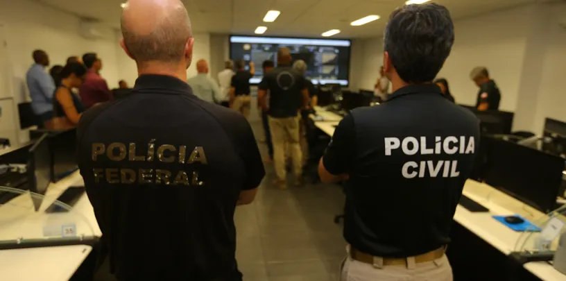 Sala de monitoramento ou treinamento com várias pessoas diante de computadores e uma tela grande ao fundo exibindo dados. Em primeiro plano, duas pessoas estão de costas vestindo camisetas pretas com os textos “POLÍCIA FEDERAL” e “POLÍCIA CIVIL”. O ambiente sugere colaboração entre forças policiais em análise ou operação institucional.