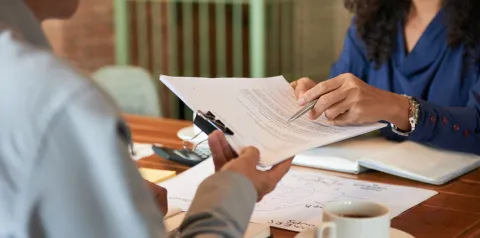 Duas pessoas analisam e trocam documentos durante uma reunião de trabalho em uma mesa de escritório