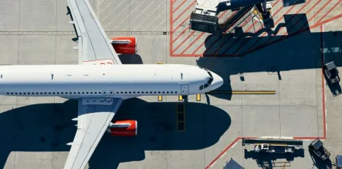 Vista aérea de avião estacionado no aeroporto, conectado à ponte de embarque e cercado por equipamentos de solo.