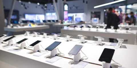 Exposição de smartphones alinhados sobre balcões em loja de eletrônicos, com dispositivos conectados a suportes de segurança e etiquetas de preço
