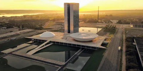Vista aérea do Congresso Nacional em Brasília ao pôr do sol, com edifícios icônicos, espelhos d’água e vias ao redor