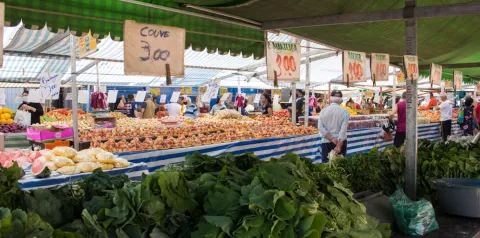 Bancas de frutas e verduras em feira ao ar livre, com produtos organizados sobre mesas e placas de preços visíveis, como ‘couve 3,00