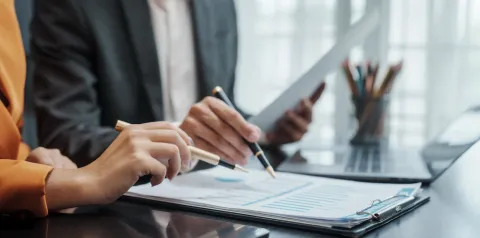 Duas pessoas analisam documentos com gráficos e relatórios impressos sobre uma mesa de escritório, utilizando canetas para apontar informações