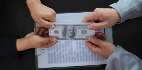 Quatro mãos de pessoas vestindo roupas formais seguram simultaneamente uma nota de 100 dólares sobre uma prancheta com documentos desfocados ao fundo. A cena sugere negociação financeira, disputa ou possível situação de suborno em ambiente corporativo.
