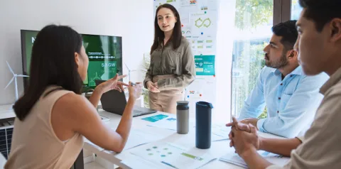 Grupo de pessoas reunidas em sala de reunião analisa materiais relacionados à sustentabilidade, enquanto uma pessoa apresenta informações em pé ao lado de um quadro com gráficos e diagramas. Sobre a mesa há documentos, um laptop e garrafas reutilizáveis, e ao fundo aparece uma tela com dados de energia limpa e turbinas eólicas, indicando discussão sobre iniciativas ESG e gestão sustentável.