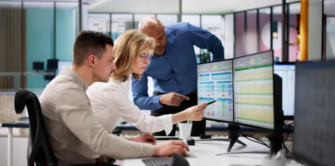 Três profissionais analisam dados em monitores de computador, apontando para planilhas e gráficos em ambiente de escritório colaborativo
