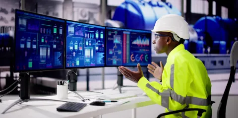 Pessoa usando capacete de segurança e roupa de alta visibilidade, sentada diante de vários monitores com gráficos e diagramas industriais. As telas exibem dados de processos, enquanto a pessoa interage gesticulando, em um ambiente que parece uma planta industrial com equipamentos ao fundo.