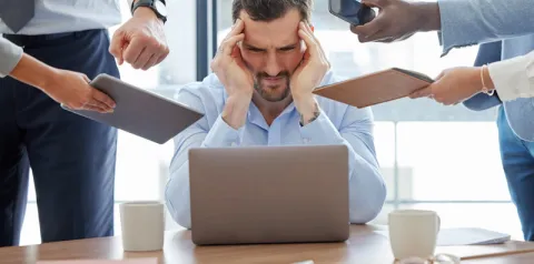 Pessoa sentada à mesa de escritório em frente a um laptop, com as mãos nas laterais da cabeça, enquanto outras pessoas ao redor estendem dispositivos como celular, tablet e bloco de anotações em sua direção. A mesa possui uma xícara e objetos de trabalho, e o ambiente é iluminado por luz natural.
