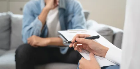 Cena de atendimento profissional em ambiente interno, onde uma pessoa segura uma caneta e um clipboard para anotações enquanto outra pessoa está sentada ao fundo em um sofá. A composição sugere uma consulta ou entrevista, com foco em coleta de informações e conversa em contexto clínico ou de aconselhamento.