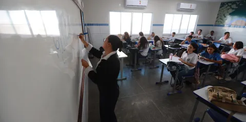 Pessoa escreve em quadro branco enquanto estudantes sentados em carteiras acompanham a aula em sala de aula iluminada