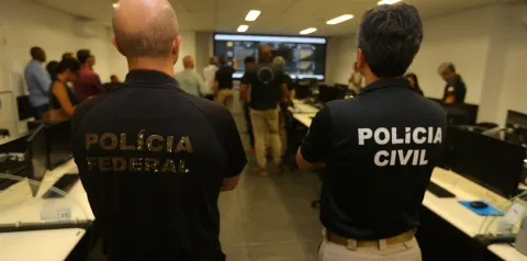 Sala de monitoramento ou treinamento com várias pessoas diante de computadores e uma tela grande ao fundo exibindo dados. Em primeiro plano, duas pessoas estão de costas vestindo camisetas pretas com os textos “POLÍCIA FEDERAL” e “POLÍCIA CIVIL”. O ambiente sugere colaboração entre forças policiais em análise ou operação institucional.