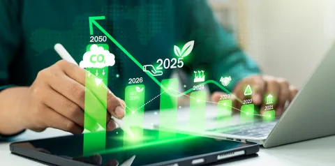 Pessoa utilizando uma caneta digital em um tablet ao lado de um laptop, enquanto gráficos verdes ascendentes e ícones sobrepostos representam metas de sustentabilidade, como redução de CO₂, neutralidade de carbono e crescimento até 2050. A cena sugere análise de dados ambientais e planejamento estratégico sustentável.
