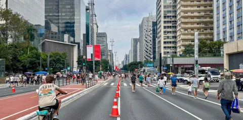 Vista da Avenida Paulista em São Paulo com trecho fechado para veículos e aberto para pedestres e ciclistas, com cones delimitando as faixas. Diversas pessoas caminham pela via entre prédios altos, enquanto um ciclista aparece em primeiro plano. Ao fundo, são visíveis edifícios comerciais, sinalização urbana e movimento intenso de pessoas ao longo da avenida.